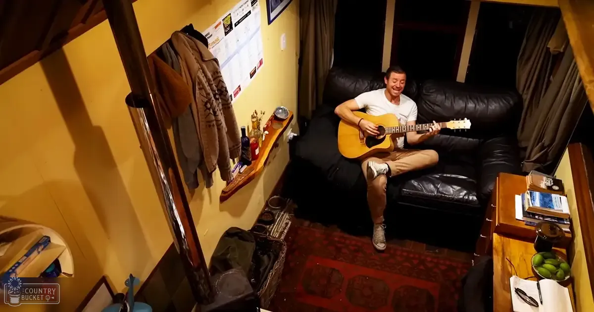 A young adult male playing guitar in a living room with yellow walls, black couch, and green fruits on a side table.