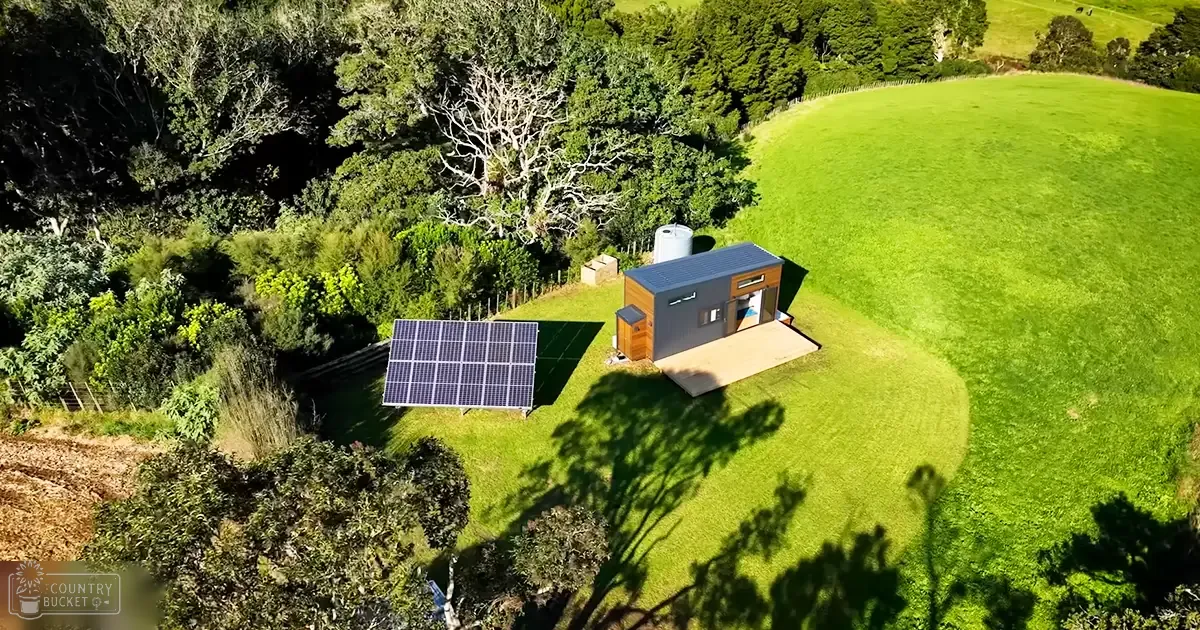 A rural scene with a small house with a gray roof and wooden accents, large solar panels, lush green grass, trees, and a water tank behind the house; peaceful countryside setting likely in spring or summer.