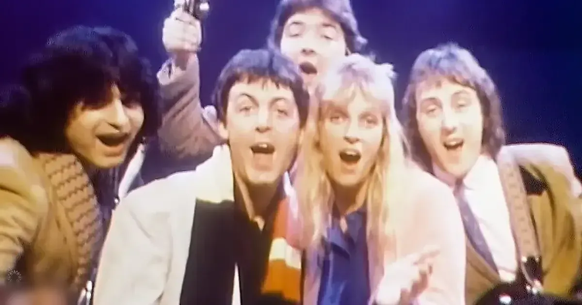 Group performance scene with adults dressed in classic late 1970s to early 1980s attire, set against a dark stage backdrop under warm lighting.