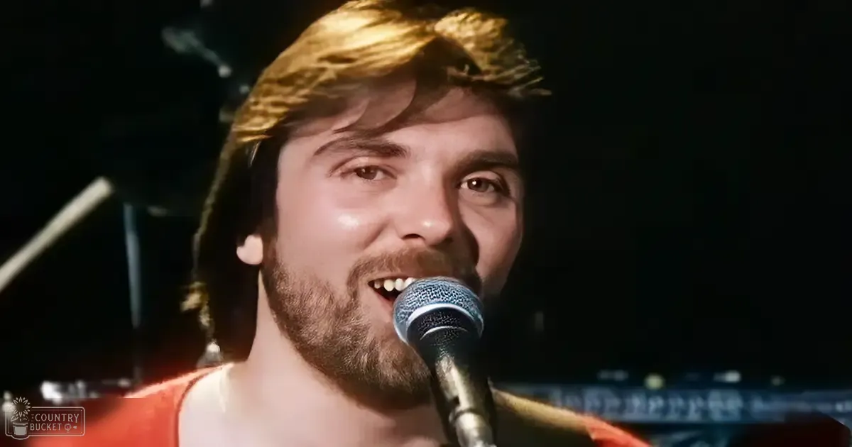 A male adult singer performing at a concert with an orange garment and white strap over the shoulder against a dark stage background.
