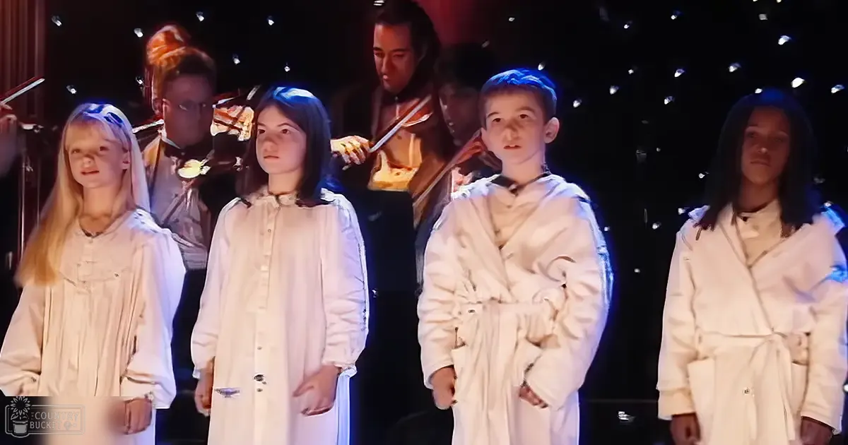 A group of children dressed in traditional choir robes perform on a stage with small decorative lights and a dark curtain backdrop.