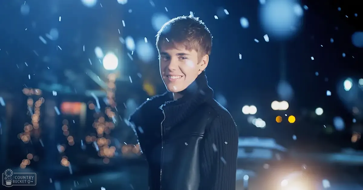 A young adult male wearing a ribbed wool turtleneck sweater stands amidst urban night snowfall with street lamps and out-of-focus buildings in the background.