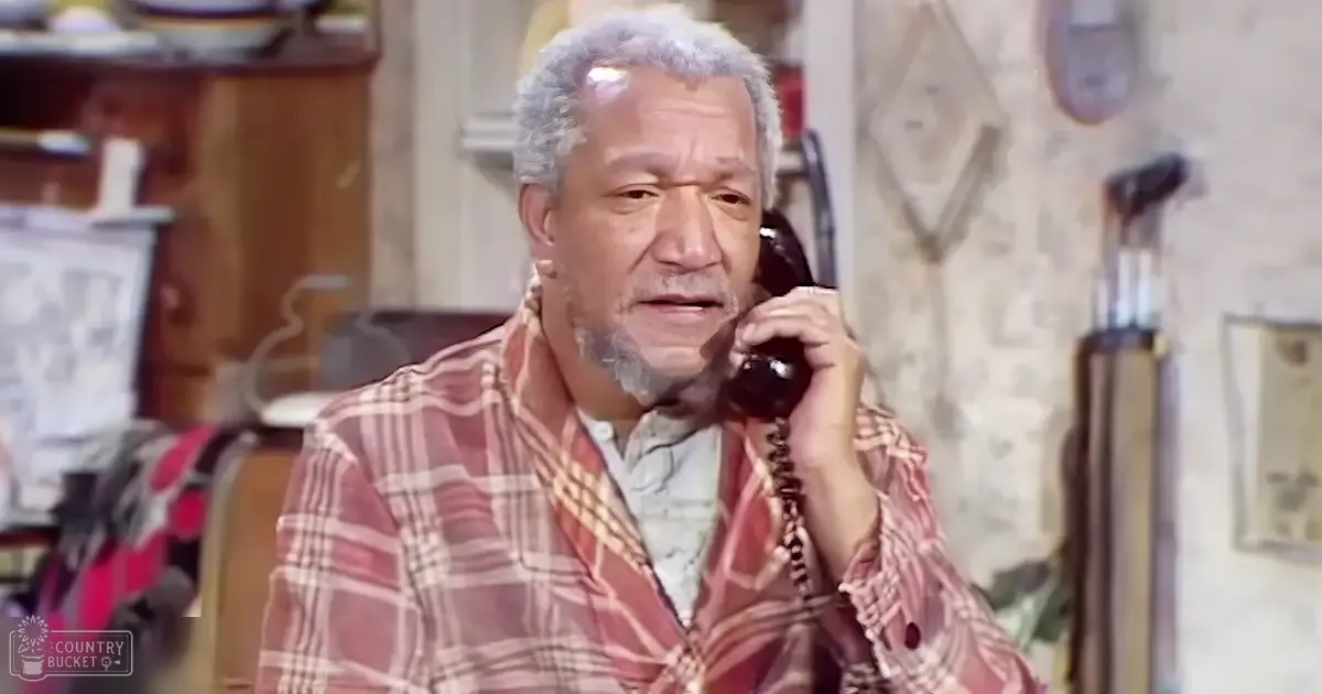 An older adult male in a plaid shirt holding an old-fashioned rotary phone in a kitchen setting with wooden cabinets and utensils, suggesting mid-20th century attire.