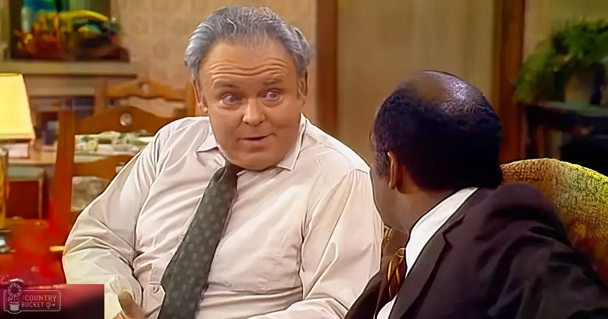 An older adult male wearing a white dress shirt with rolled-up sleeves, dark tie with small white dots, and light-colored suit jacket sits in a living room with another adult male seated to his right. The setting includes wooden furniture, a lamp with a yellow shade, plants, and decorative items on a shelf.