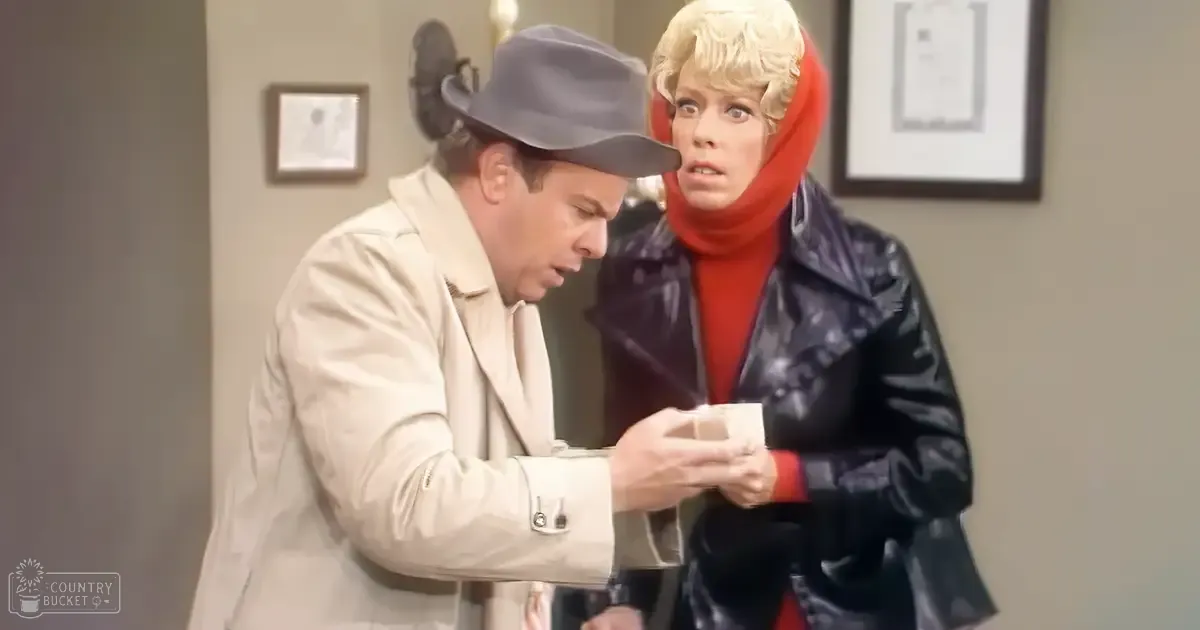 An adult male singer wearing a light beige trench coat with a gray fedora-style hat, engaged in conversation with an older female in a dark blue leather jacket and red turtleneck sweater within a professional indoor setting.