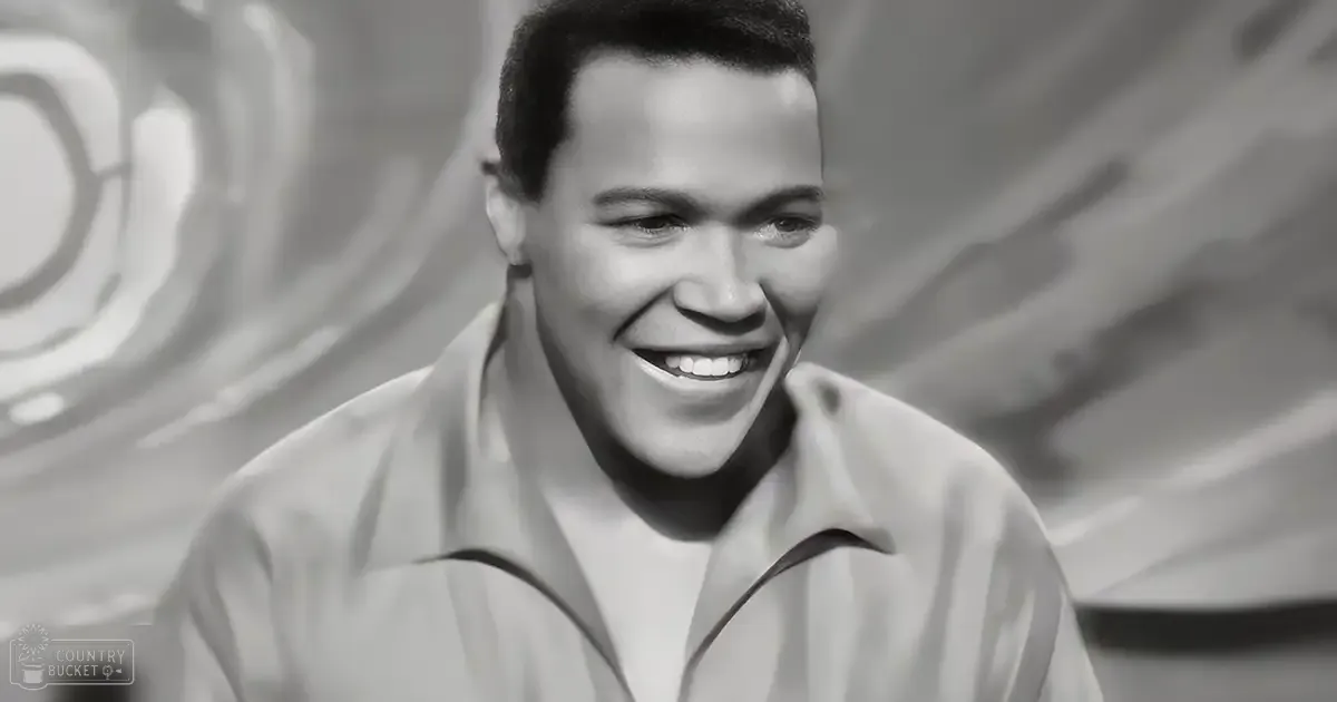 A young adult male stands indoors, wearing a light-colored collared shirt with relaxed fit and long sleeves.
