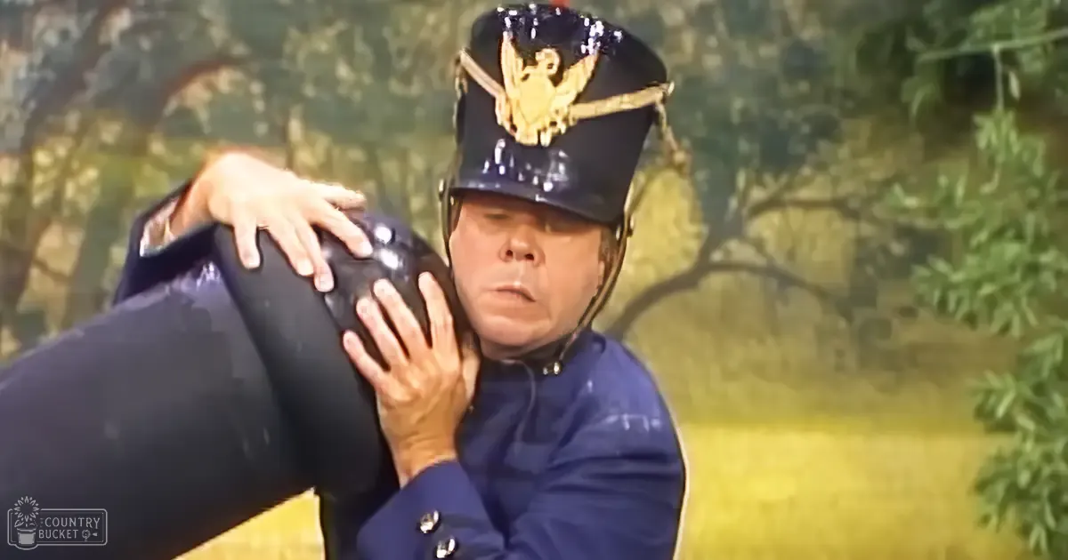 Middle-aged adult male in a dark blue military uniform outdoors amidst greenery with trees and natural light illuminating the scene.