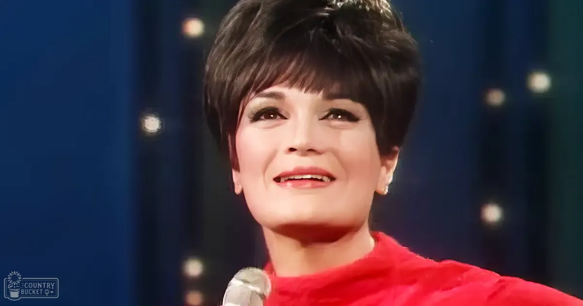 A middle-aged female in a red, shiny formal dress from mid-20th century indoors with stage lighting and microphone.