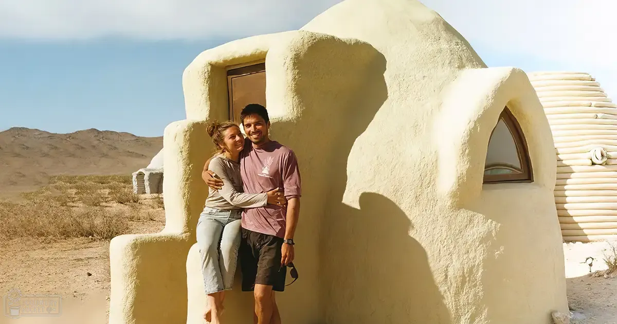 A young adult female with light brown hair tied back, wearing a long-sleeved gray shirt and blue jeans stands next to an adult male in a desert setting. The adobe structure behind them suggests their home or shelter.