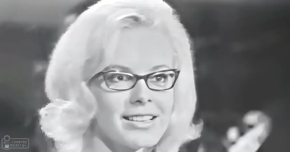 An older adult female with short white hair in a studio setting, wearing black-framed glasses and a formal garment from the mid-20th century.