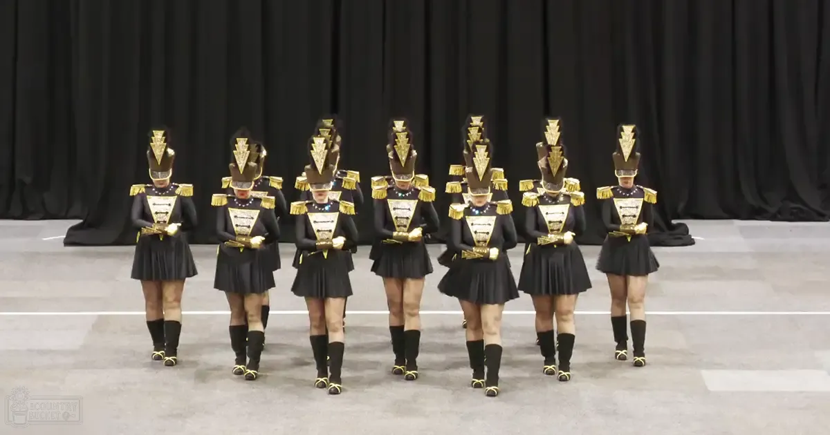 Young adult female performers in a dance or marching band setting, wearing black pleated skirts with gold trim, black tops with gold details, black knee-high socks and boots, ornate headgear, holding small instruments against a plain indoor arena backdrop.