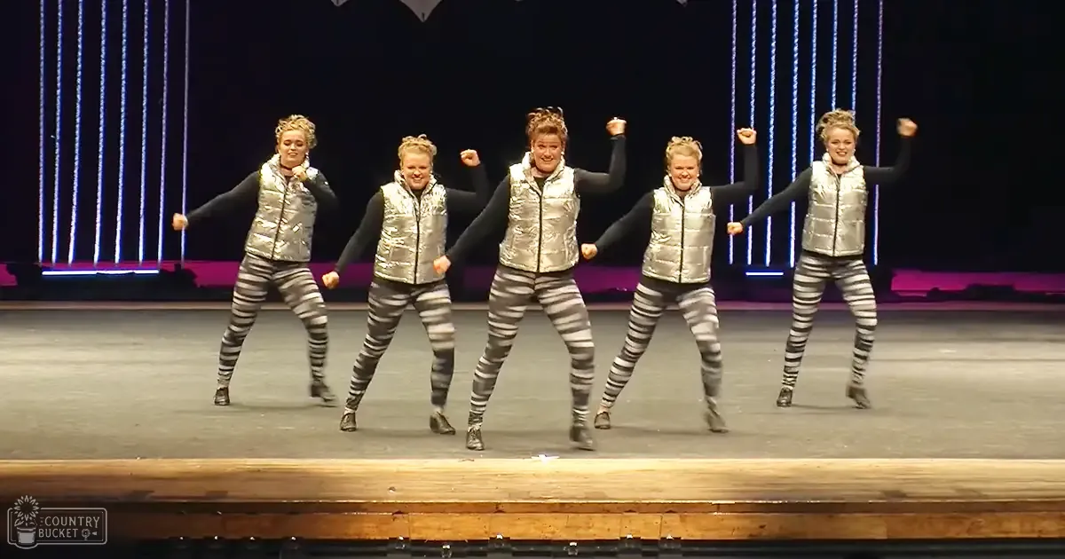 A group of five dancers in shiny silver vests over black tops, striped leggings, and black boots performing on a dark indoor stage with patterned lighting.