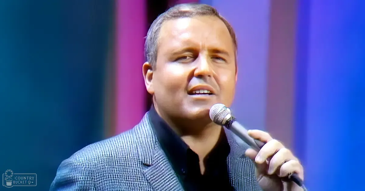 A middle-aged adult male speaks into a microphone at an indoor event, wearing a dark blue collared shirt with a dark gray checkered blazer.