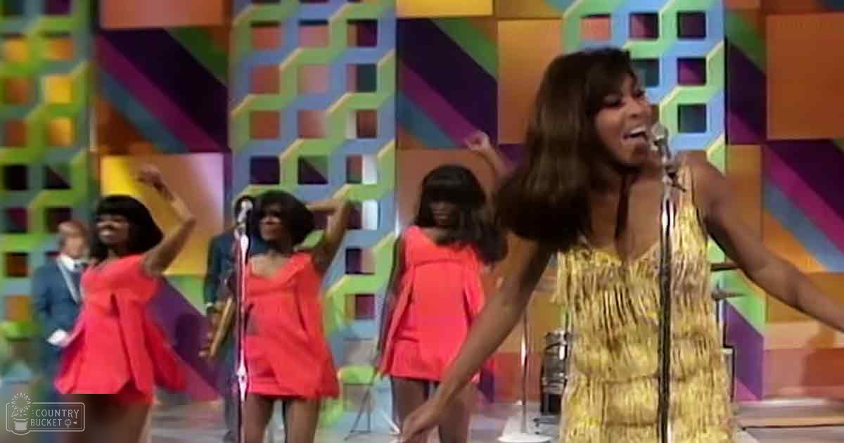 A female performer in a gold fringe dress with backup dancers wearing bright red dresses on stage under vivid 1960s-inspired lighting.