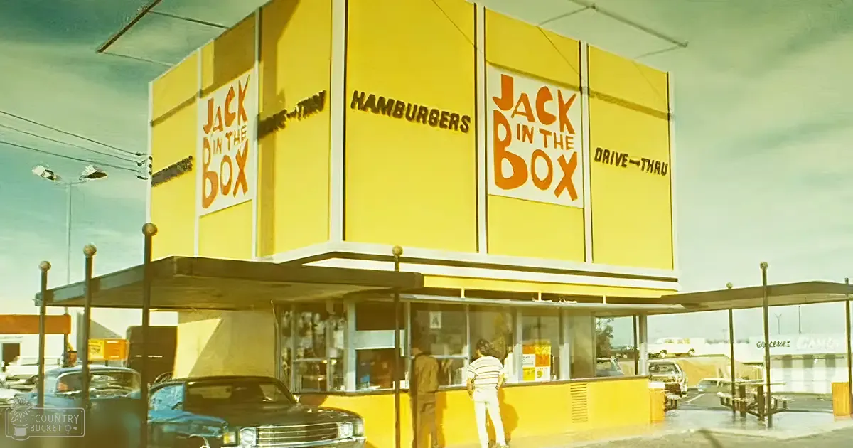 Urban Jack in the Box restaurant scene, clear skies, daytime, picnic tables, yellow building, blue sky, various shades of blue and white cars, brown and beige pavement.