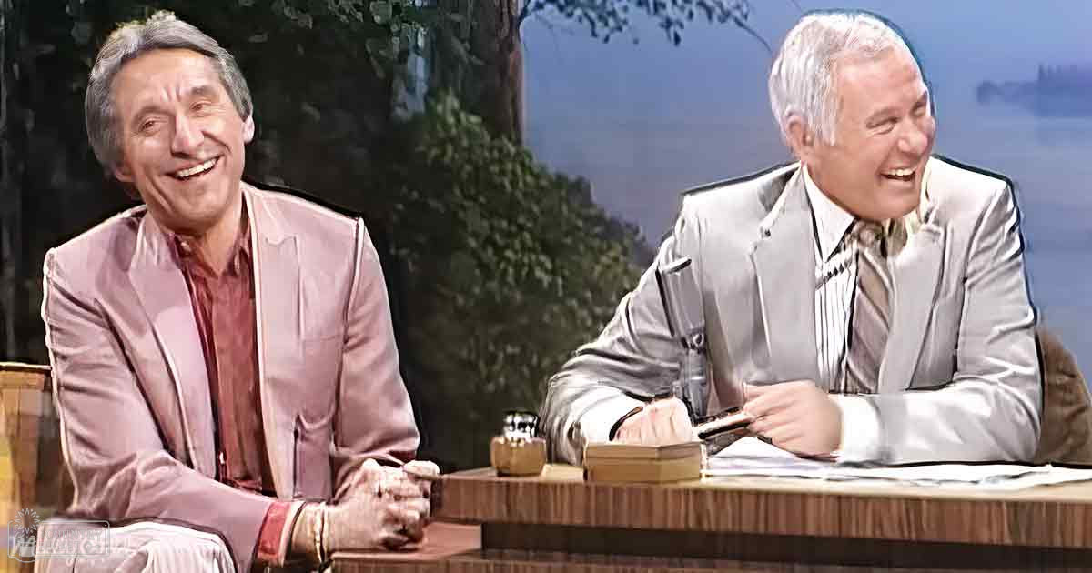Two older adult males, one wearing a light pink blazer and maroon shirt, the other in a light gray suit jacket over a striped white collared shirt, seated at a wooden desk with papers and a small container, against a backdrop of an outdoor scene viewed through windows.
