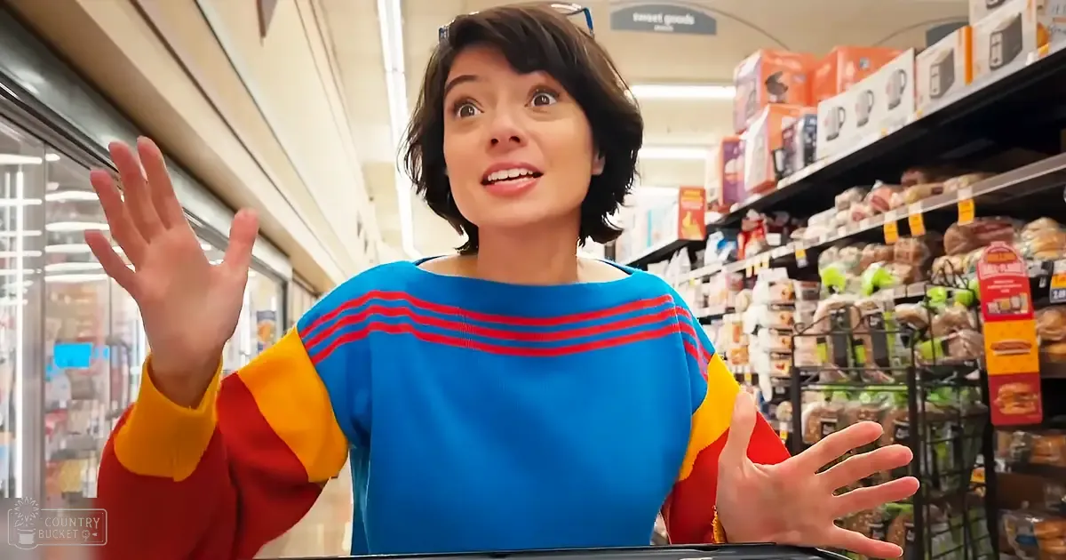 A young adult female wearing a relaxed blue sweater with red and yellow color blocks in a well-lit grocery store aisle filled with various products.