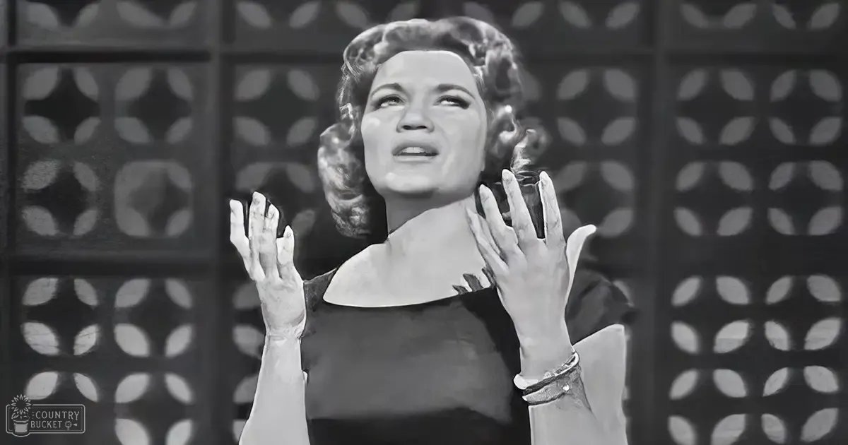 A middle-aged female in a formal, mid-20th-century-style black dress indoors with decorative metalwork on patterned walls and diffused lighting.