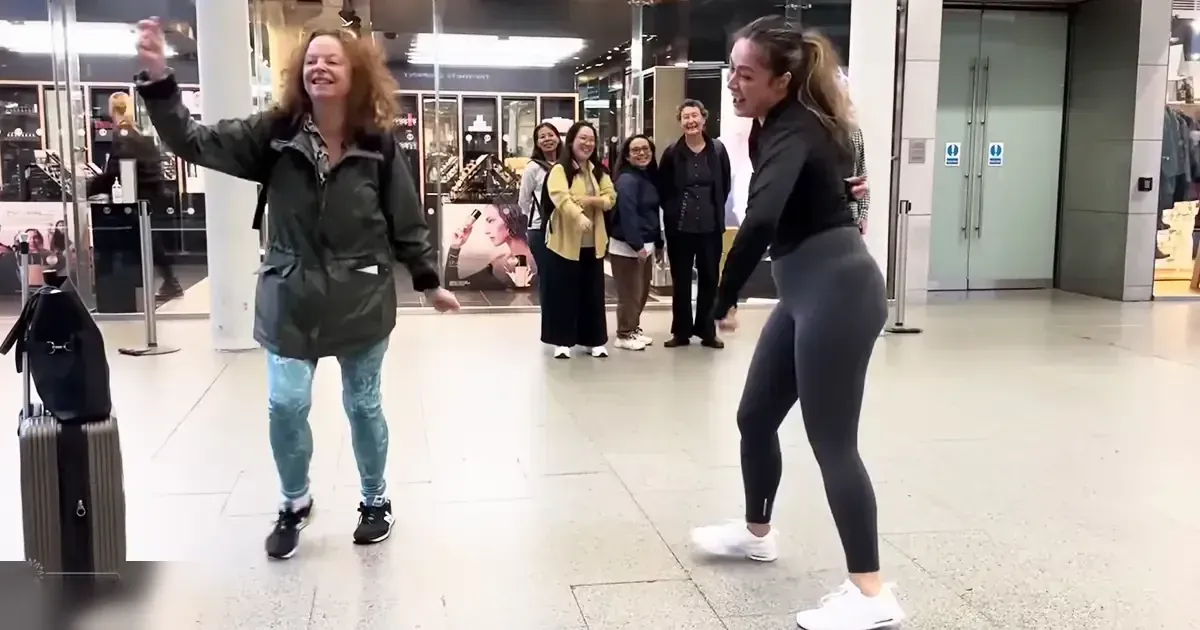 Two young adult females dancing in an indoor public space, possibly an airport terminal; casual attire and a line of onlookers standing nearby.