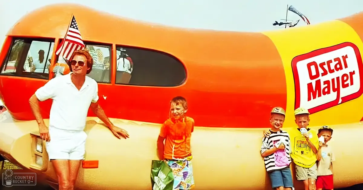 A man with middle-aged appearance and sunglasses poses in front of an Oscar Mayer Wienermobile, surrounded by three children wearing colorful outfits at a fair or festival under clear blue skies.