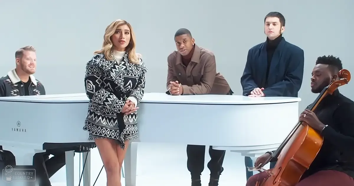 A young adult female stands in front of a white grand piano, with other individuals seated behind her. The setting is an indoor studio or performance space.