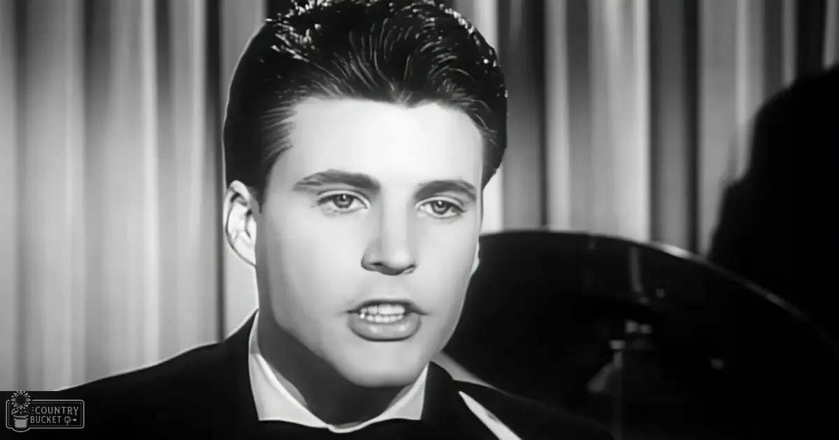 A young adult male in a tailored formal suit jacket and white collared shirt stands indoors, possibly on a stage with a piano visible behind him. The setting has neutral tones from curtains or drapes.