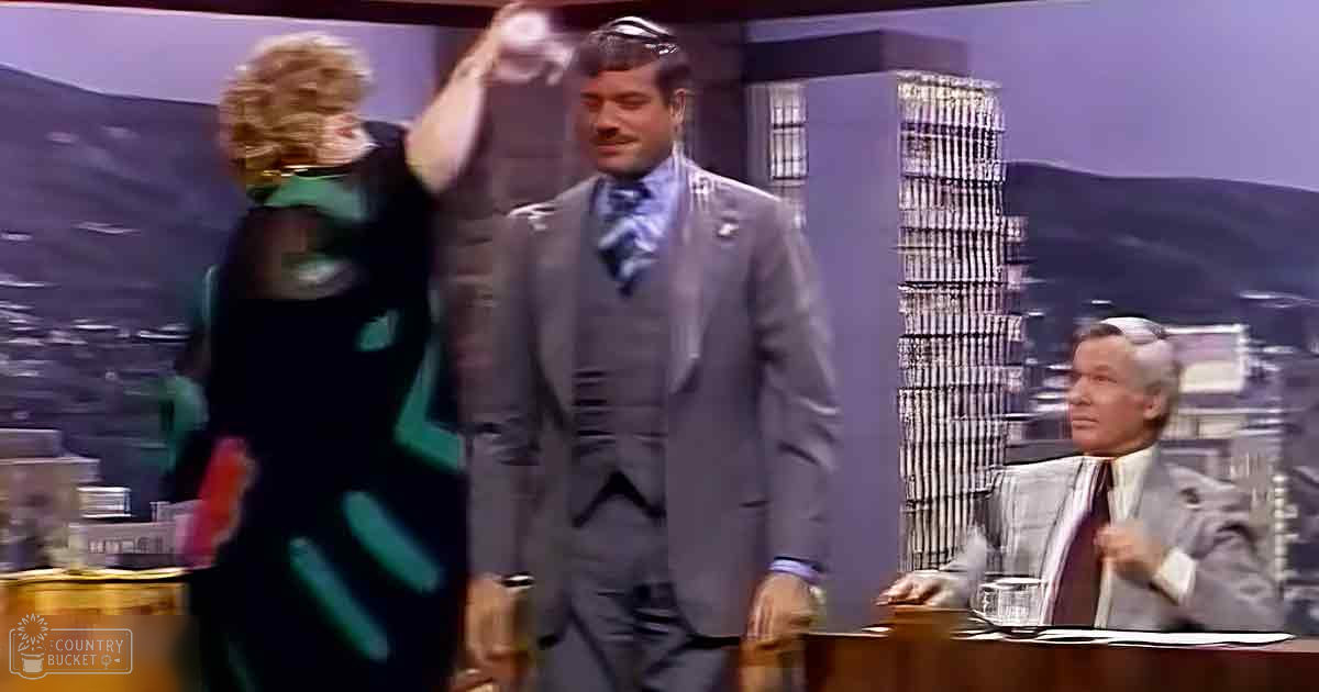 Adult male in a gray three-piece suit with a white shirt and blue patterned tie, standing indoors against a cityscape backdrop; adult female wearing a dark green dress with an abstract geometric pattern, seated nearby. The scene suggests a professional or entertainment industry setting from the mid-20th century.
