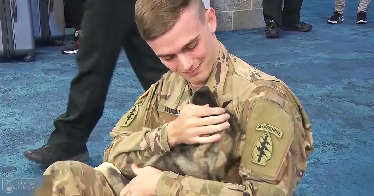 Young adult male soldier in a military uniform holding a small German Shepherd mix puppy indoors, possibly at an airport or event hall.
