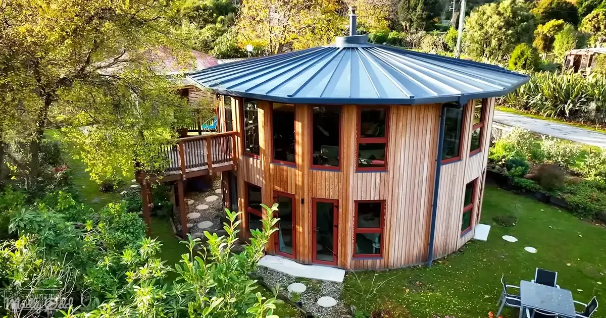 A unique octagonal house with a blue metal roof nestled in a lush garden setting on a sunny afternoon.