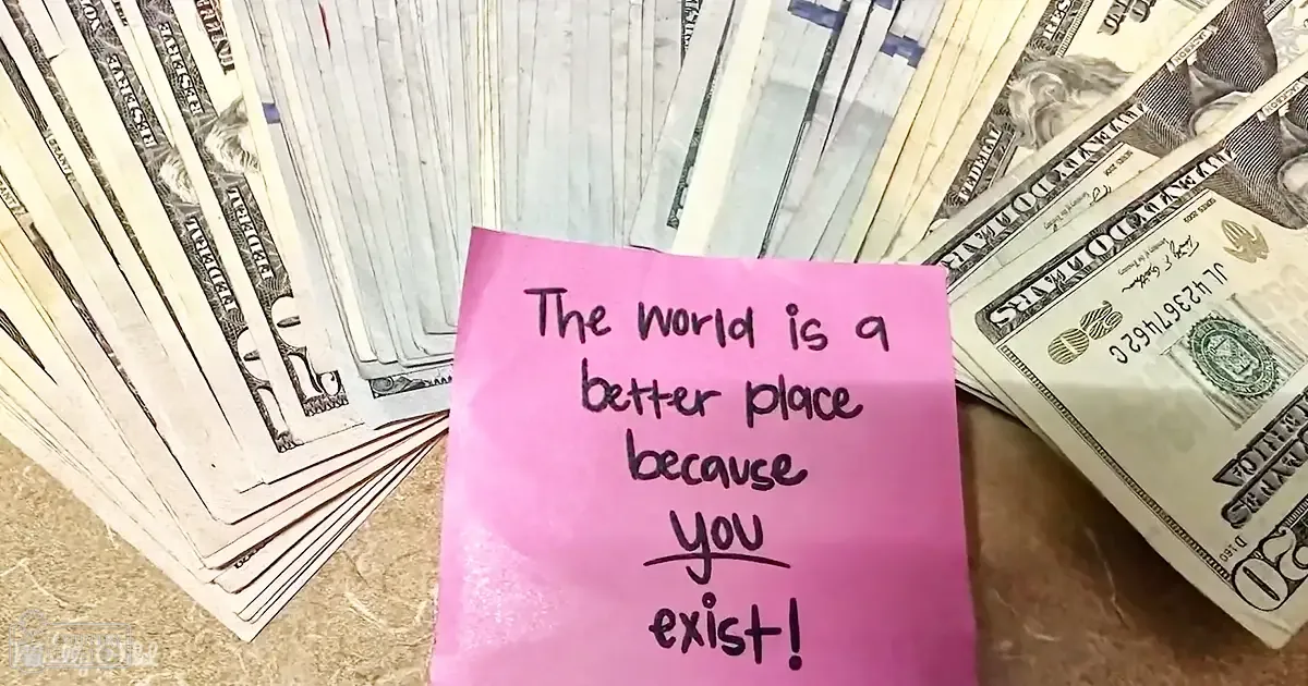 Pink sticky note with inspirational text among US dollar bills on a wooden surface indoors.