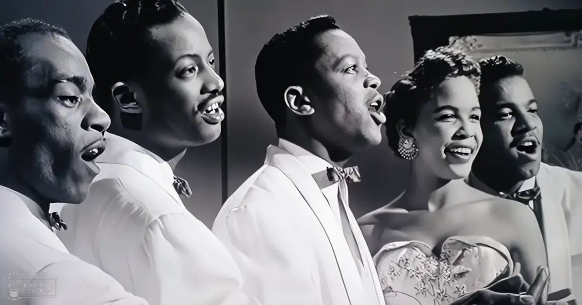 A group of adult male and female performers singing in a mid-20th-century studio setting, dressed formally with intricate patterns and accessories.