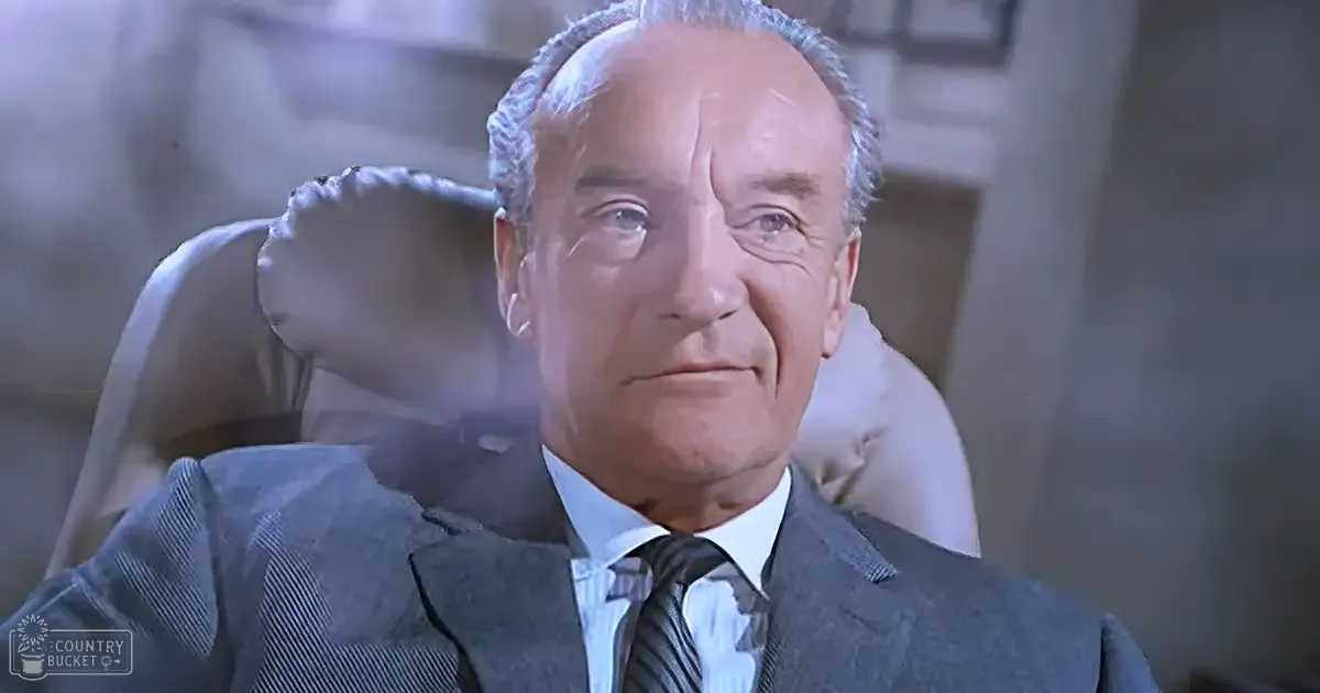 An older adult male in a formal setting, wearing a dark gray suit jacket with subtle pinstripes and a striped tie.