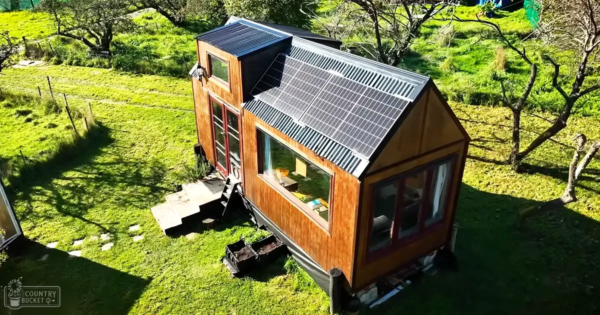 A small wooden house with solar panels in a rural setting surrounded by grassy fields and trees under clear daylight conditions.