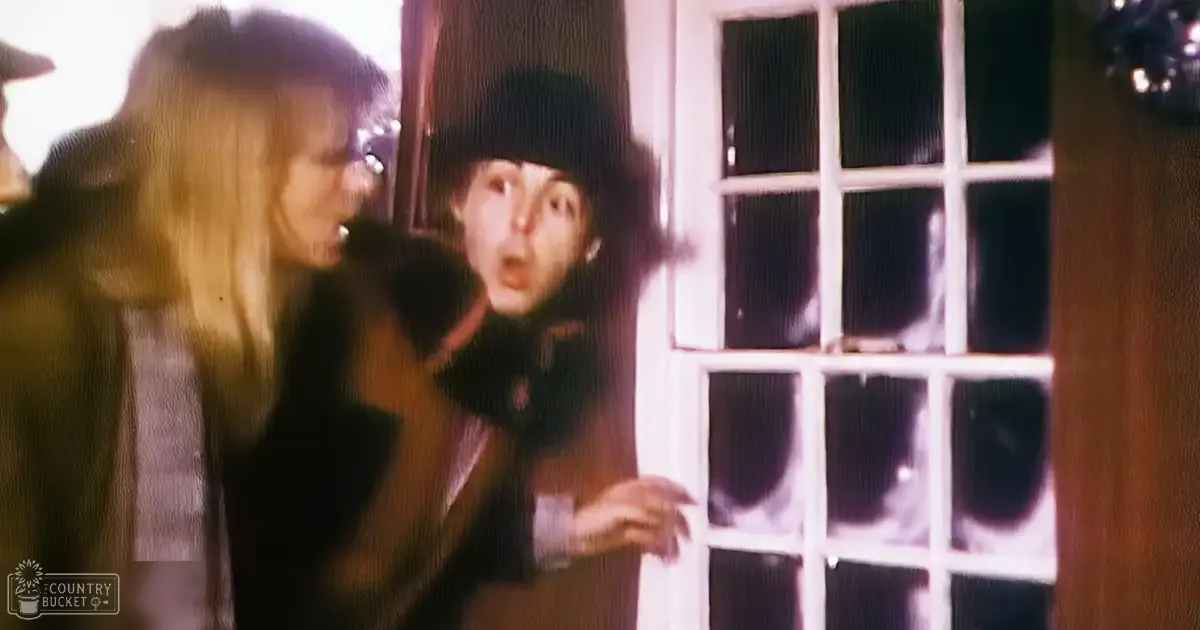 An adult male singer wearing a black hat, dark jacket with high collar, scarf or turtleneck under the jacket; an adult female in a dark coat and light shirt underneath stands to his left. White door with multiple glass panes highlights the setting.