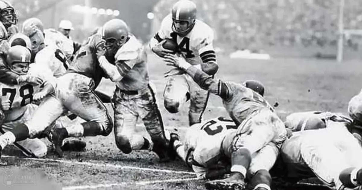 A football game in progress at an outdoor stadium with players wearing white jerseys, dark pants, and cleats on a field marked by yard lines and goalposts.