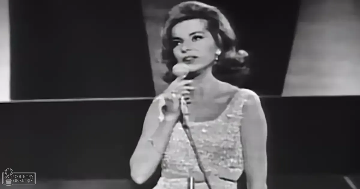 An adult female singer in a studio setting wearing an elegant sleeveless dress with intricate detailing, holding a microphone and pearl earrings.