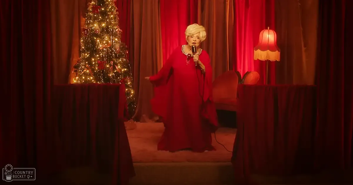 An older adult female performer wearing a long red mid-20th century gown with flared sleeves and high-collared neckline, standing against a red curtain backdrop with a Christmas tree in the background on an indoor stage or performance space at night.