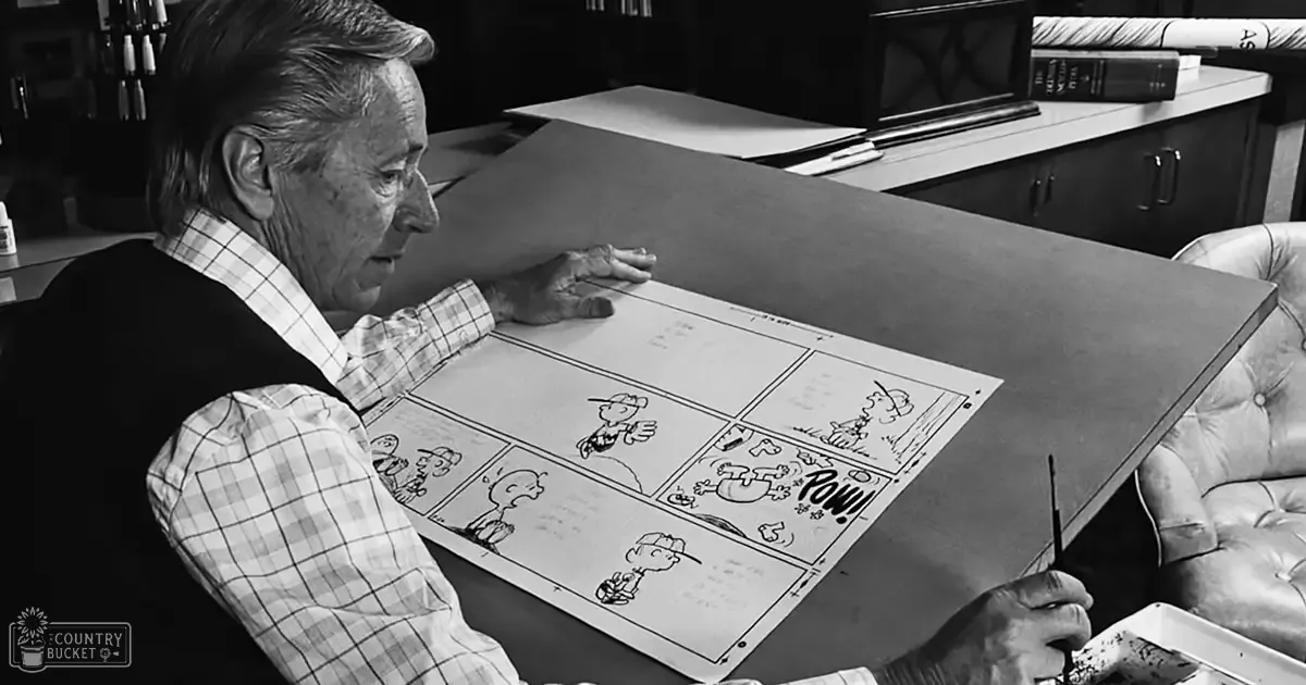 An older adult male working on comic strips in a mid-20th century office setting with checkered shirt and dark vest, surrounded by books and an office chair, black and white image.