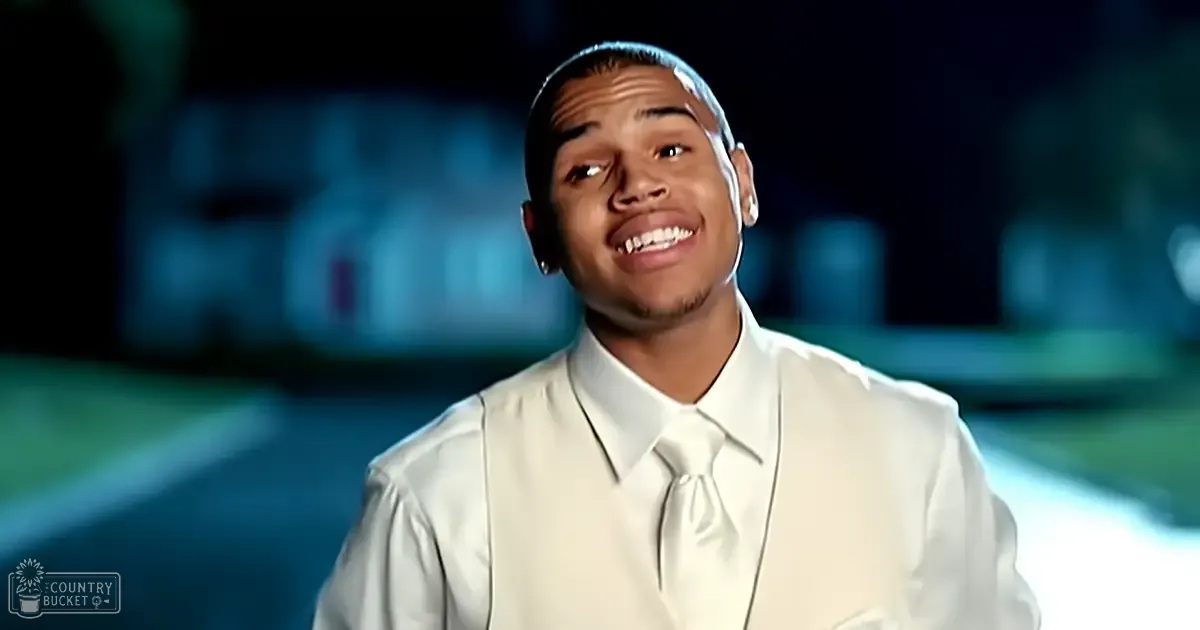A young adult male stands outdoors at night, wearing a formal white dress shirt with a white tie and vest, against a backdrop of blurred greenery under artificial light.