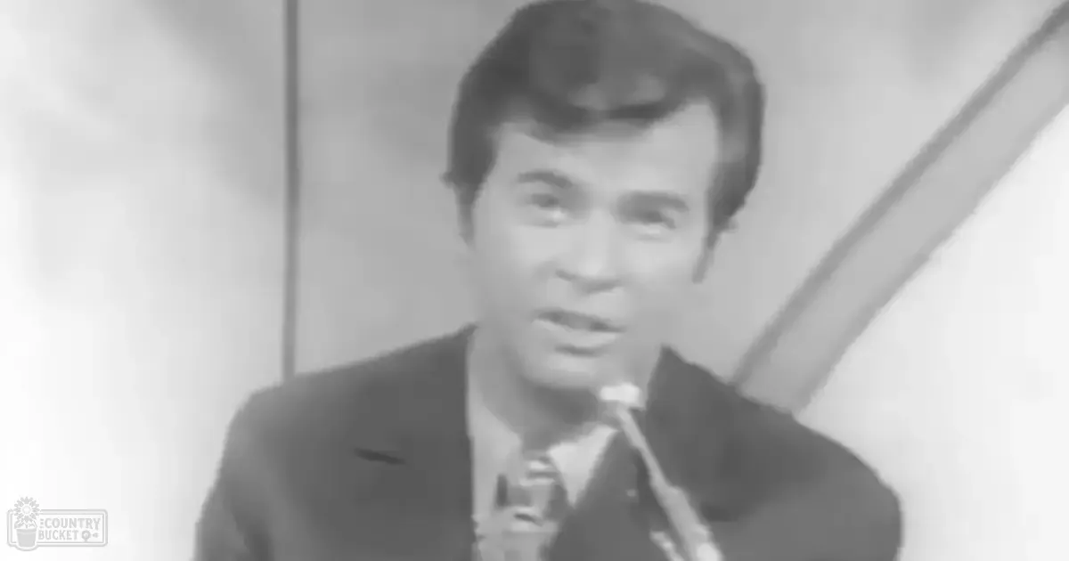 Young adult male in formal suit holding microphone indoors with plain walls as background.