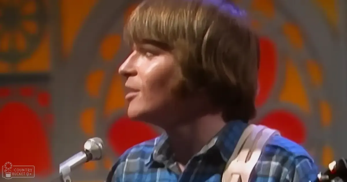 A young adult male wearing a relaxed-fit plaid shirt with short sleeves, standing against a colorful patterned wall in what appears to be a recording studio or performance space.