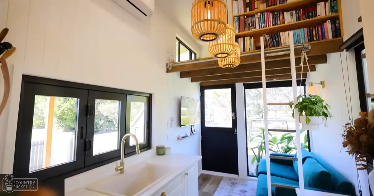 A modern, cozy interior space with wooden bookshelves filled with books, a hanging basket light fixture, and a blue couch in a residential setting with natural daylight streaming through windows showing greenery outside.