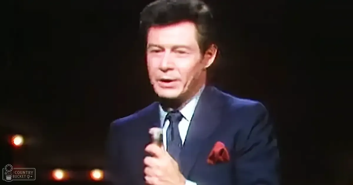 A middle-aged male singer in a dark blue suit with a white shirt and red pocket square, holding a microphone on stage under dim lighting.