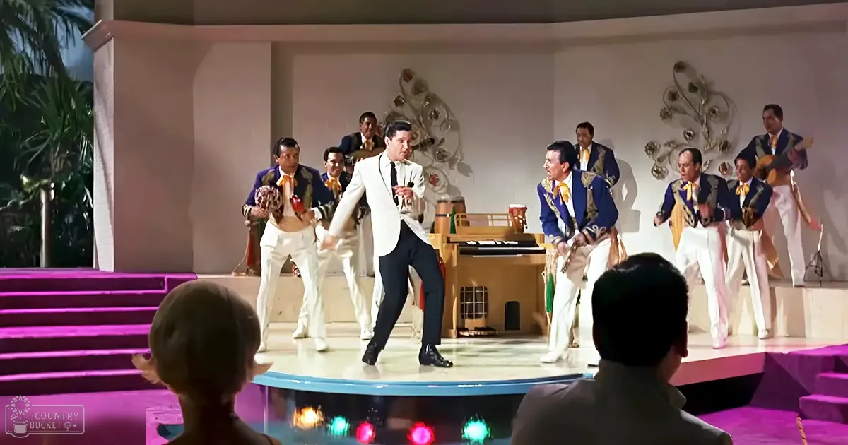 An adult male singer dancing in a white tuxedo with a mariachi band dressed in traditional attire and formal shoes on stage, with a piano, decorative wall art, and purple carpeted steps leading up to the stage. The setting suggests an evening or night scene indoors at a hotel lobby or resort.