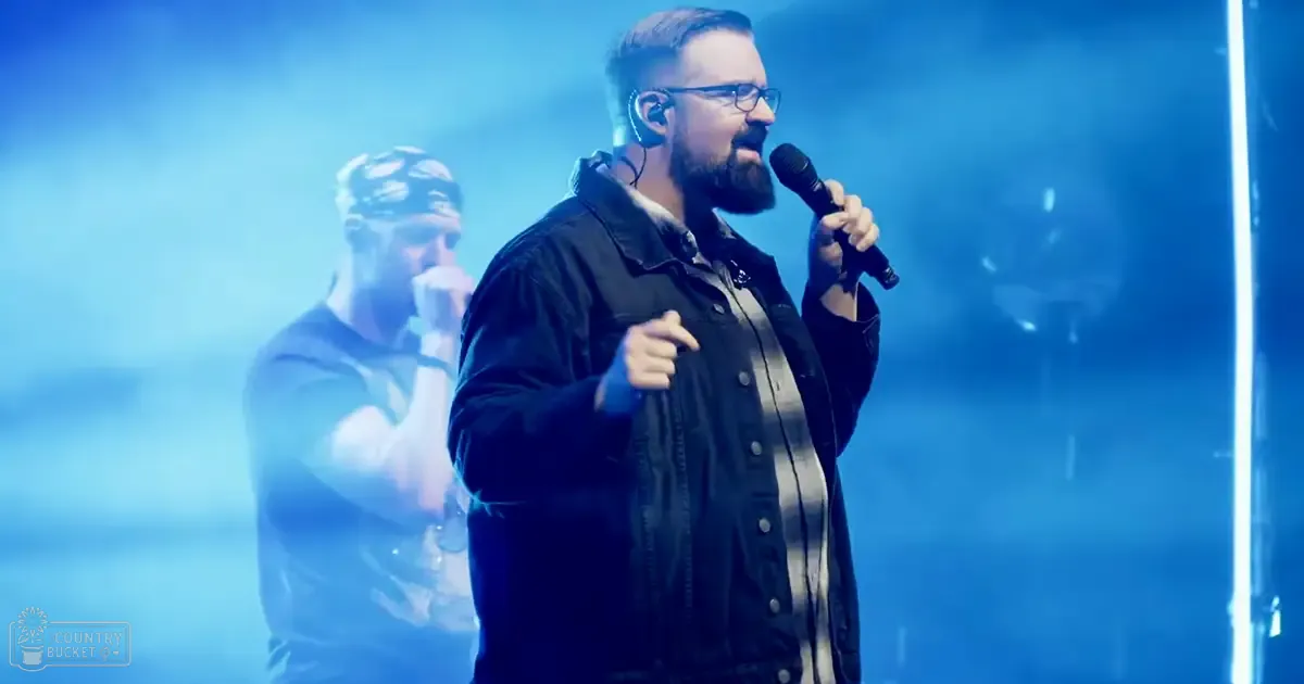 Adult male performer wearing a dark jacket with light-colored stripes, glasses, and earpiece microphone; another adult male in the background wears a patterned shirt and headband, possibly holding a microphone. Indoor stage setting with blue lighting creating dramatic atmosphere.