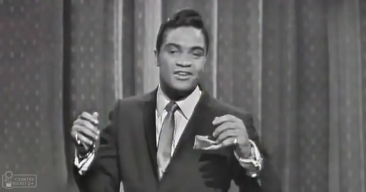 A young adult male in a formal suit with patterned tie, standing against a backdrop with vertical stripes, possibly indicating an indoor performance or presentation setting.
