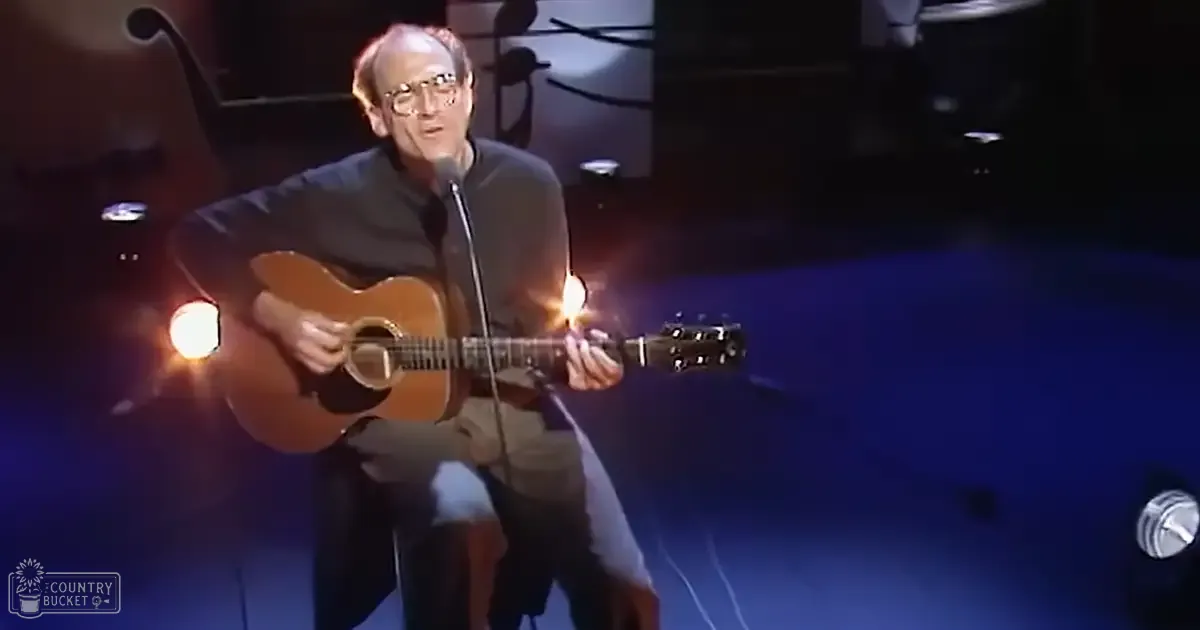 A middle-aged man wearing a dark gray long-sleeved shirt, playing an acoustic guitar on stage with visible 'Madly Odd' text in the corner.