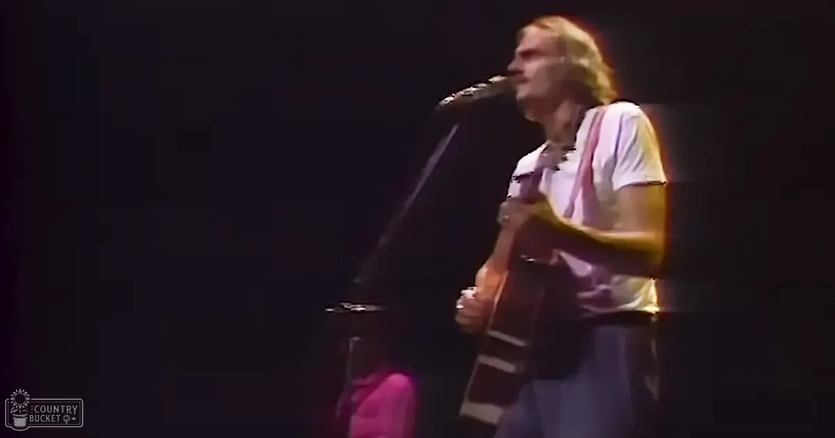 A male musician, likely young adult, performs on stage wearing a white T-shirt with pink and purple stripes at a concert.