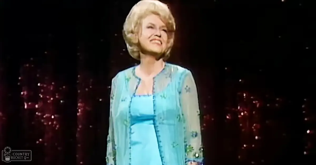 An older adult woman in a stage setting, wearing a light blue floral dress and matching jacket with dark curtains and sparkling lights behind her.