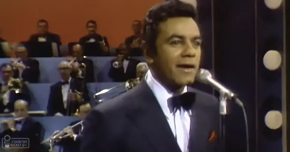 Adult male performer in dark suit and bow tie on stage with seated audience, brass instruments visible, evening indoor setting under artificial light.